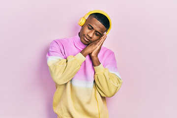 Young black man listening to music wearing headphones sleeping tired dreaming and posing with hands together while smiling with closed eyes.