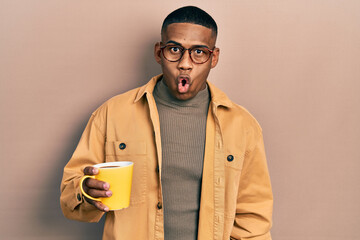 Young black man wearing glasses and drinking a cup of coffee scared and amazed with open mouth for...