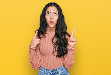 Hispanic teenager girl with dental braces wearing casual clothes amazed and surprised looking up and pointing with fingers and raised arms.