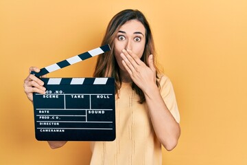 Young hispanic girl holding video film clapboard covering mouth with hand, shocked and afraid for...