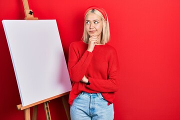 Beautiful blonde woman standing by painter easel stand thinking concentrated about doubt with...