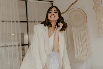 Charming brunette curly woman in great mood poses in white cozy room. Pretty young lady in stylish...
