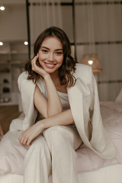 Brown-eyed woman in beige pants and linen jacket smiles widely and looks into camera. Brunette curly lady in suit sits on soft bed.