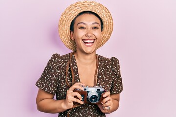 Beautiful hispanic woman with short hair wearing summer hat holding vintage camera smiling and laughing hard out loud because funny crazy joke.