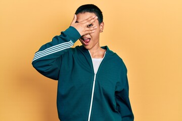 Beautiful hispanic woman with short hair wearing sporty jacket peeking in shock covering face and eyes with hand, looking through fingers with embarrassed expression.