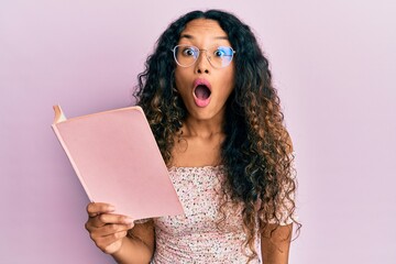 Young latin woman reading a book with glasses scared and amazed with open mouth for surprise,...