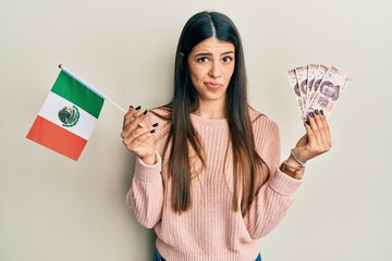 Young hispanic woman holding mexico flag and mexican pesos banknotes skeptic and nervous, frowning...