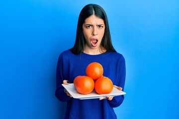 Young hispanic woman holding plate with fresh oranges clueless and confused expression. doubt...