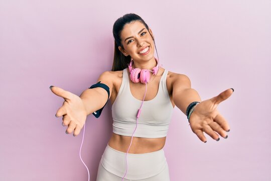Young hispanic woman wearing gym clothes and using headphones smiling cheerful offering hands giving assistance and acceptance.