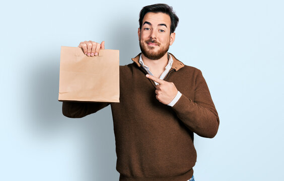 Young hispanic man holding take away paper bag smiling happy pointing with hand and finger