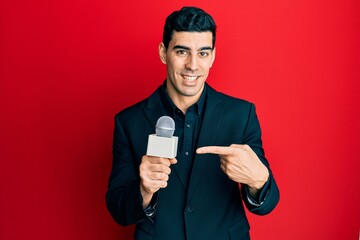 Handsome hispanic man holding reporter microphone smiling happy pointing with hand and finger