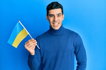 Handsome hispanic man wearing ukraine flag looking positive and happy standing and smiling with a confident smile showing teeth