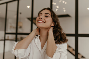 Calm happy brunette woman in cotton white shirt touches neck and smiles. Close up portrait of curly lady in light blouse poses at home.