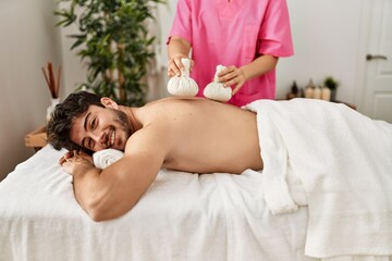 Young hispanic man smiling happy reciving herbal pouches thai massage at beauty center.