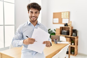 Young hispanic businessman smiling happy holding paperwork standing at the office.