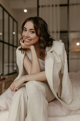 Happy brunette Caucasian woman in beige linen jacket and stylish pants sits on soft bed. Attractive curly girl in suit smiles and leans on knees.
