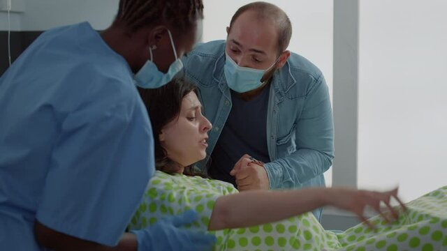Young woman in labor giving birth to baby in hospital ward with support from husband and african american obstetrics nurse. Pregnant patient having contractions pushing for delivery