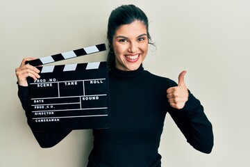 Young hispanic woman holding video film clapboard smiling happy and positive, thumb up doing...
