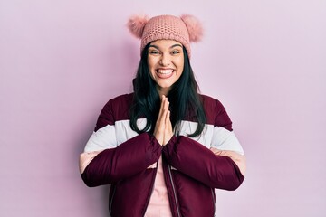 Young hispanic woman wearing wool winter sweater and cap praying with hands together asking for forgiveness smiling confident.