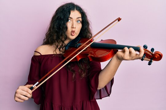 Young brunette musician woman with curly hair playing violin making fish face with mouth and squinting eyes, crazy and comical.