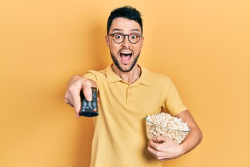 Young hispanic man eating popcorn using tv control celebrating crazy and amazed for success with...