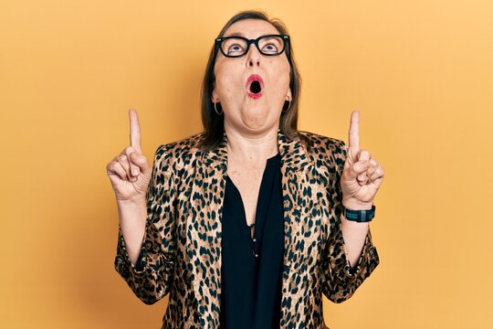 Middle age hispanic woman wearing business clothes and glasses amazed and surprised looking up and pointing with fingers and raised arms.