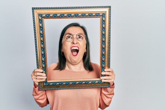 Middle age hispanic woman holding empty frame angry and mad screaming frustrated and furious, shouting with anger looking up.
