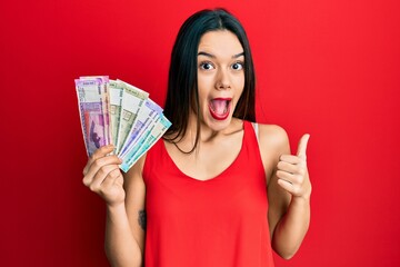 Young hispanic girl holding indian rupee banknotes pointing thumb up to the side smiling happy with open mouth