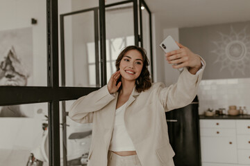 Fototapeta na wymiar Brown-eyed brunette curly woman in beige loose jacket and linen pants smiles sincerely, holds phone and takes selfie at kitchen.