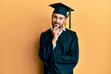 Young hispanic man wearing graduation cap and ceremony robe looking confident at the camera with...