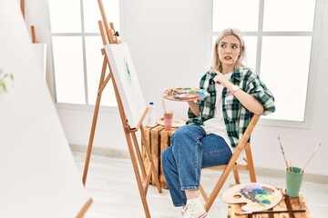 Young artist woman painting on canvas at art studio pointing aside worried and nervous with...