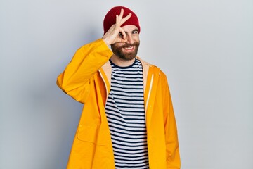 Caucasian man with beard wearing yellow raincoat doing ok gesture with hand smiling, eye looking through fingers with happy face.