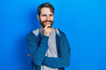 Caucasian man with beard wearing casual sweatshirt looking confident at the camera with smile with crossed arms and hand raised on chin. thinking positive.