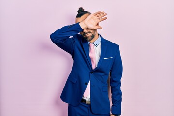 Young hispanic man wearing business suit and tie covering eyes with arm, looking serious and sad. sightless, hiding and rejection concept