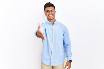 Young hispanic man wearing business shirt standing over isolated background smiling friendly offering handshake as greeting and welcoming. successful business.