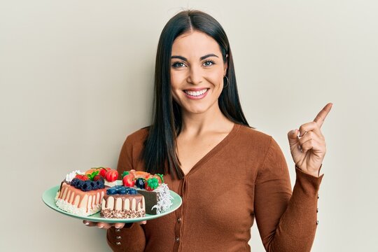 Young brunette woman holding plate with cake slices smiling happy pointing with hand and finger to the side