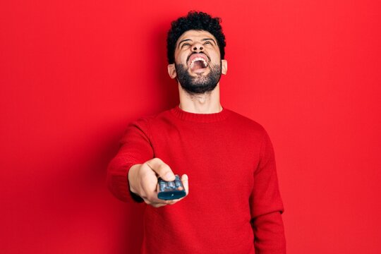Young arab man with beard holding television remote control angry and mad screaming frustrated and furious, shouting with anger looking up.