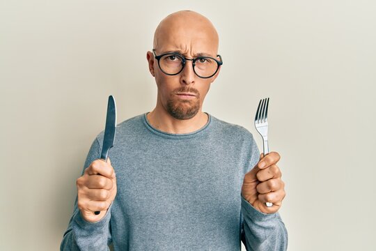 Bald man with beard holding fork and knife ready to eat skeptic and nervous, frowning upset because of problem. negative person.