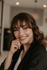 Close-up portrait of tanned attractive brunette curly woman in black stylish jacket smiles sincerely and touches cheek.