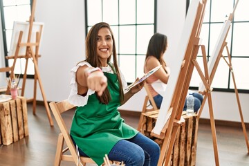 Young hispanic artist women painting on canvas at art studio smiling with happy face looking and pointing to the side with thumb up.
