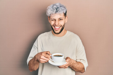 Young hispanic man with modern dyed hair drinking a cup of coffee winking looking at the camera...