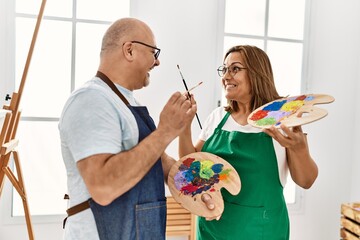 Middle age hispanic painter couple smiling happy holding paintbrush and palette at art studio.