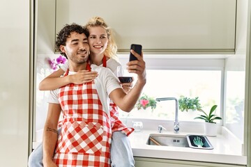 Young couple smiling happy hugging and sitting on counter. Drinking red wine and using smartphone at kitchen.