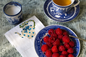 High angle selective focus view of raspberries on blue plate plate, with teacup, embroidered tea towel and sugar bowl in soft focus background