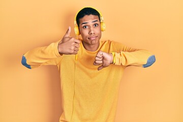 Young african american guy listening to music using headphones doing thumbs up and down,...