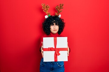 Young middle east woman wearing deer christmas hat holding gift skeptic and nervous, frowning upset because of problem. negative person.