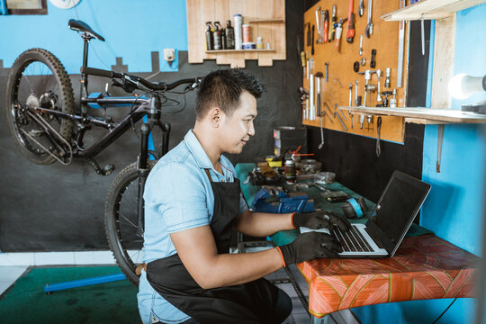 a bicycle mechanic in an apron wearing gloves sitting while using a laptop to look for spare parts while working in a workshop - Powered by Adobe