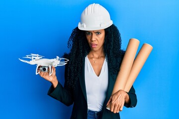 Middle age african american woman wearing safety helmet holding blueprints and drone in shock face,...