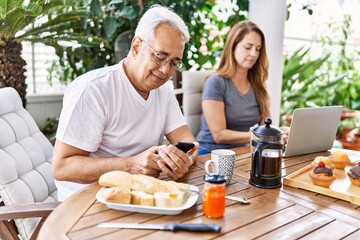 Middle age hispanic couple having breakfast using smartphone and laptop at the terrace.