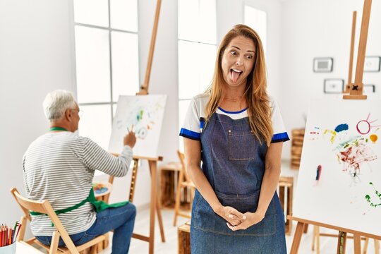 Hispanic woman wearing apron at art studio sticking tongue out happy with funny expression. emotion concept.